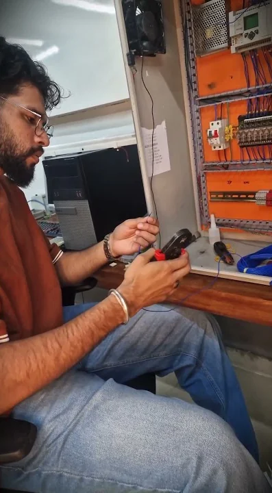 A learner holding a clamp meter next to an open orange control panel with wiring, a breaker and terminal blocks