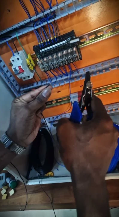 Hands using wire strippers on blue wires at a training panel with a breaker and terminal blocks