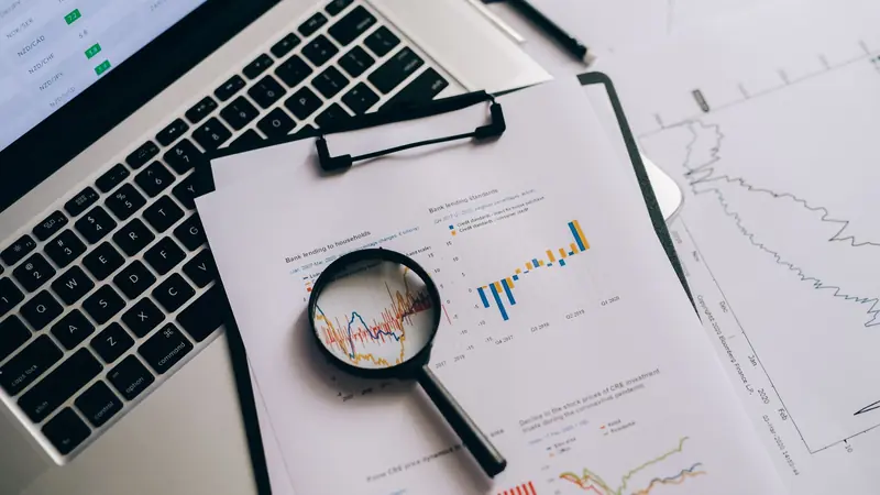 A magnifying glass over printed financial charts beside a laptop keyboard.