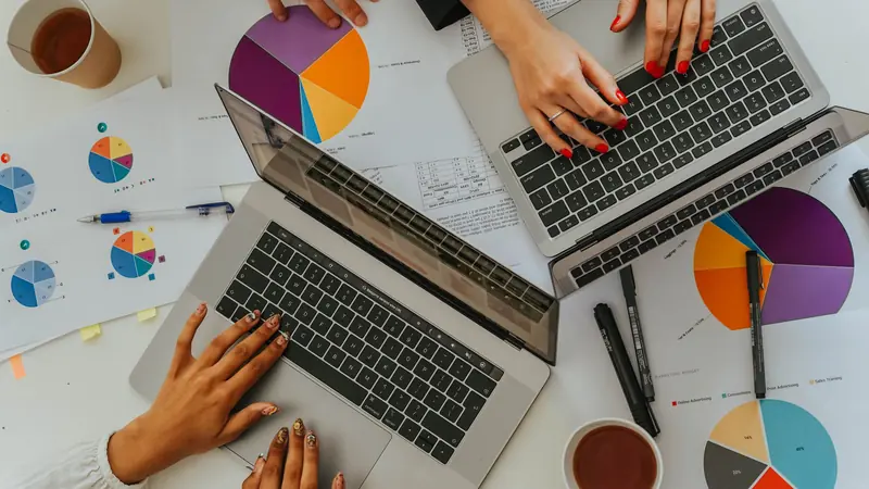 Two people at laptops over a table covered with pie charts and printed results.
