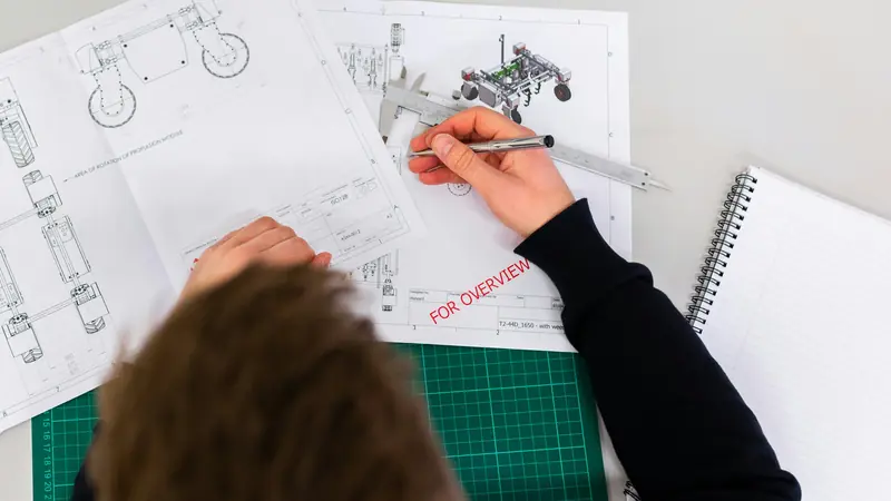 Hands marking up technical engineering drawings on a desk, seen from above.