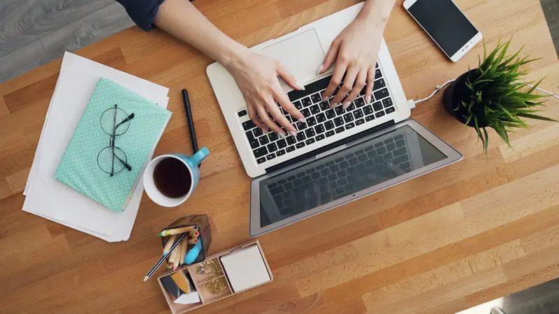 Hands typing on a laptop at a wooden desk with a notebook, glasses and coffee.
