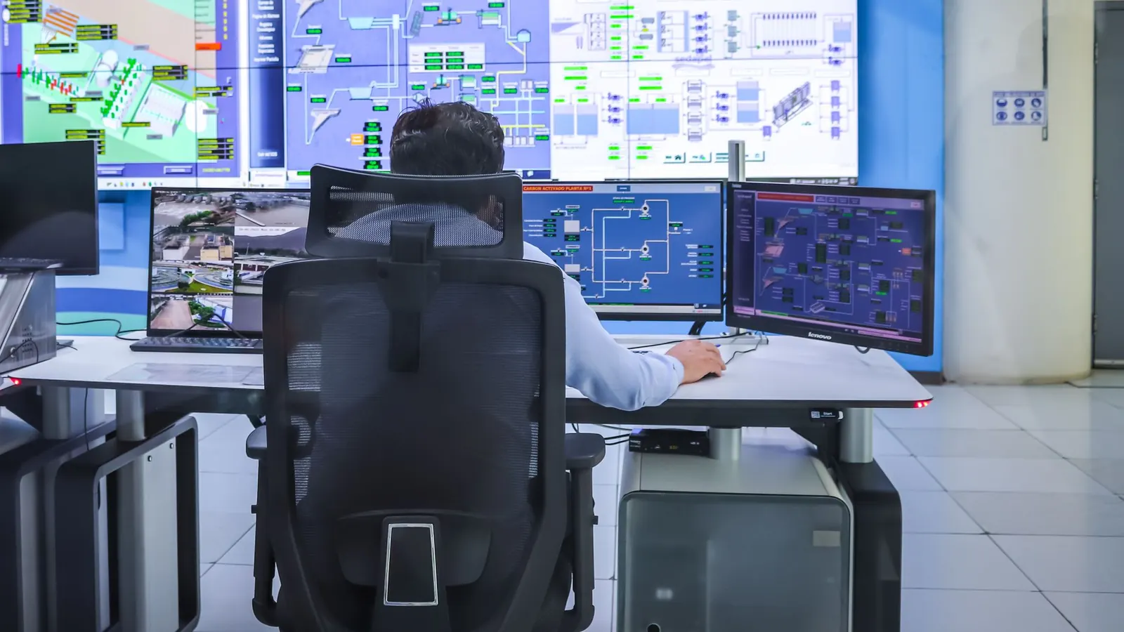 An operator at a control desk facing a video wall of process screens and trends.