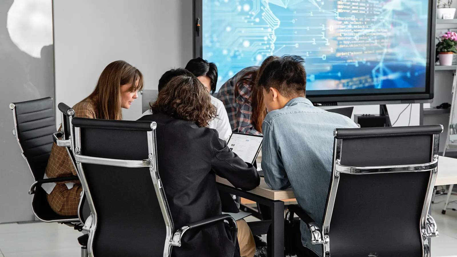 A team around a table in an office, working with laptops and a large screen.